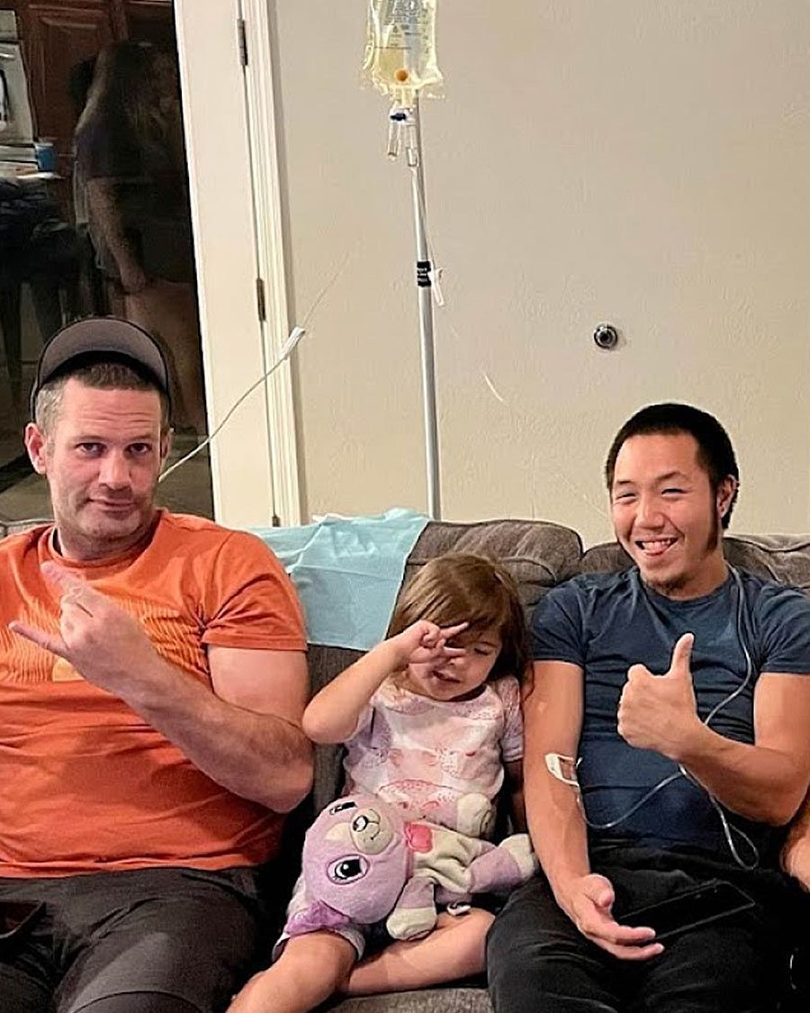 Two men receiving at-home IV infusions while sitting on a couch next to a young girl holding a stuffed animal. One man gives a thumbs up.