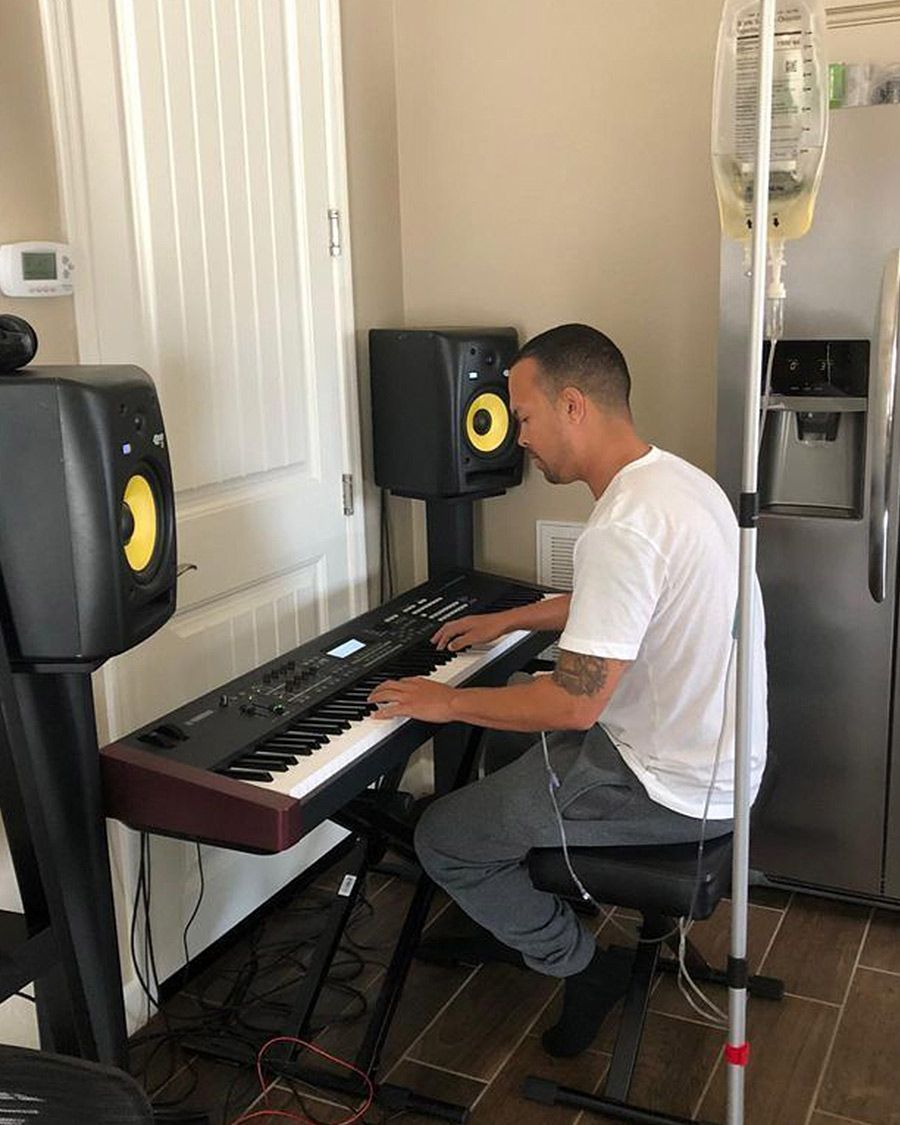 Man receiving an at-home IV therapy infusion while sitting and playing a digital keyboard in a music setup with monitor speakers.