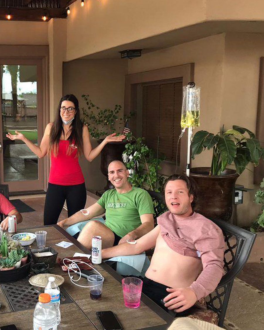 Group of friends, including two men receiving IV therapy, gathered at a patio table outdoors with a female attendant nearby.