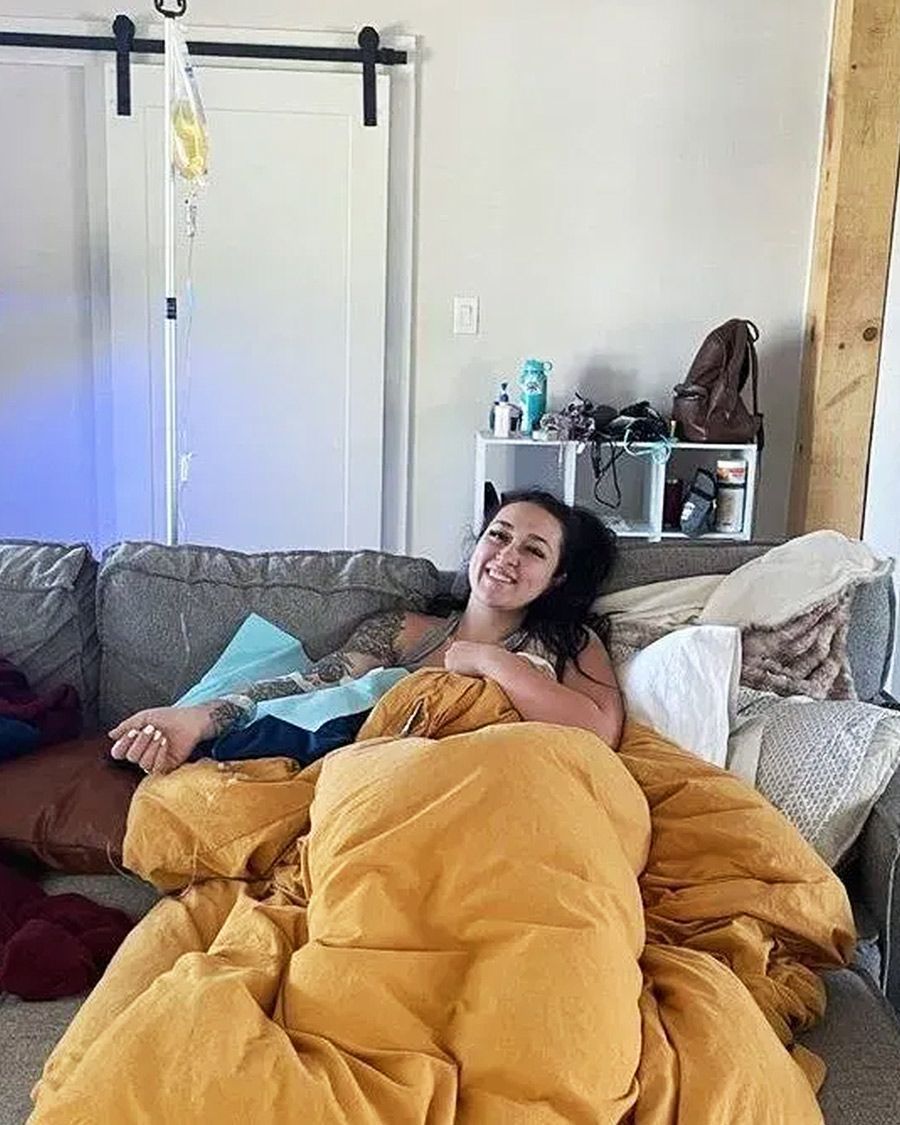 Smiling woman relaxed under a large yellow blanket on a couch, comfortably receiving an at-home IV therapy infusion.