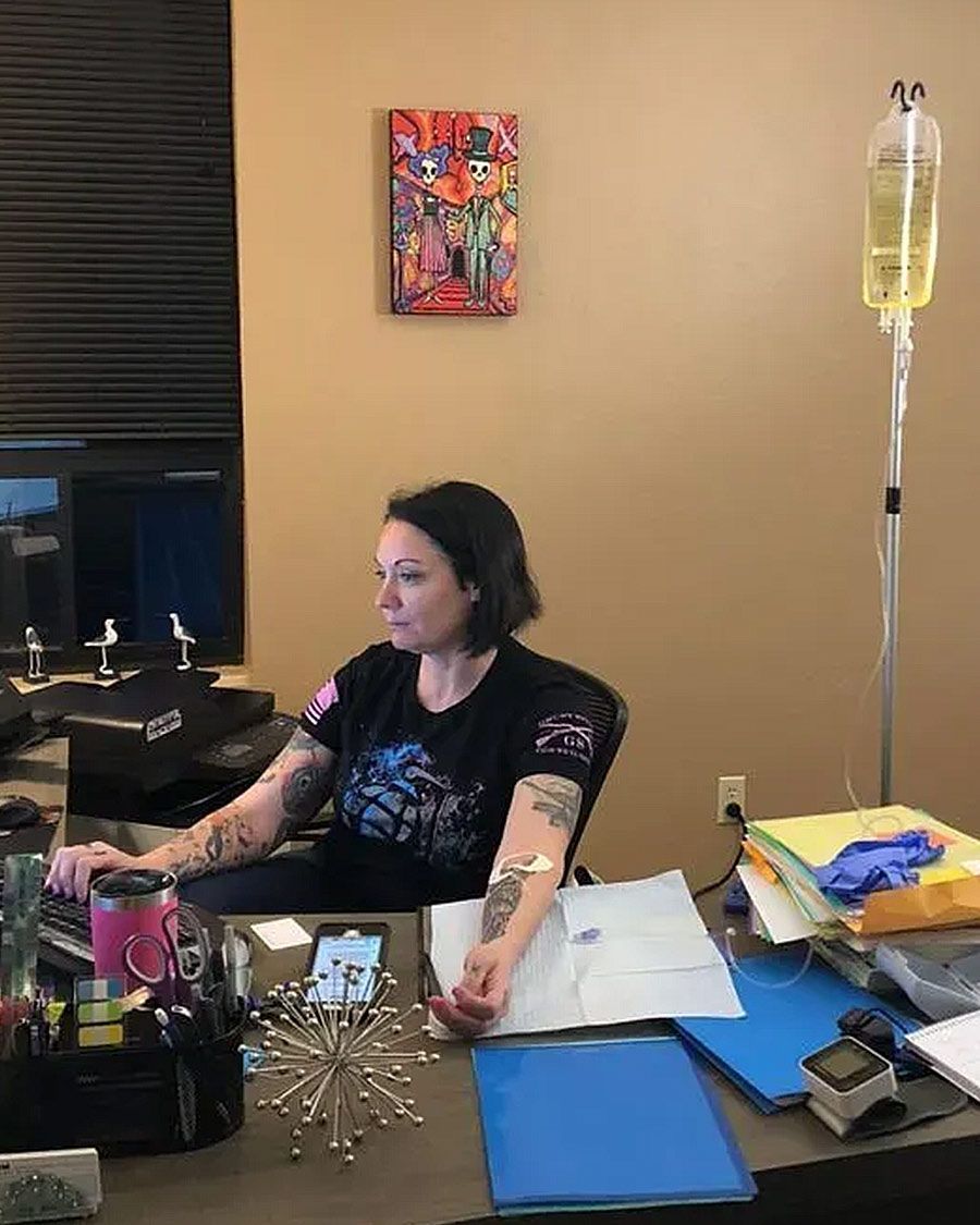 Woman with visible tattoos sitting at her cluttered home office desk, working on her computer while receiving an IV therapy infusion.