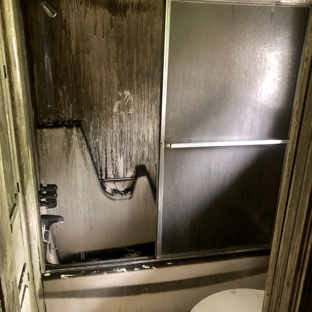 A heavily soot-stained, fire-damaged shower stall with sliding glass doors in a dark, neglected bathroom.