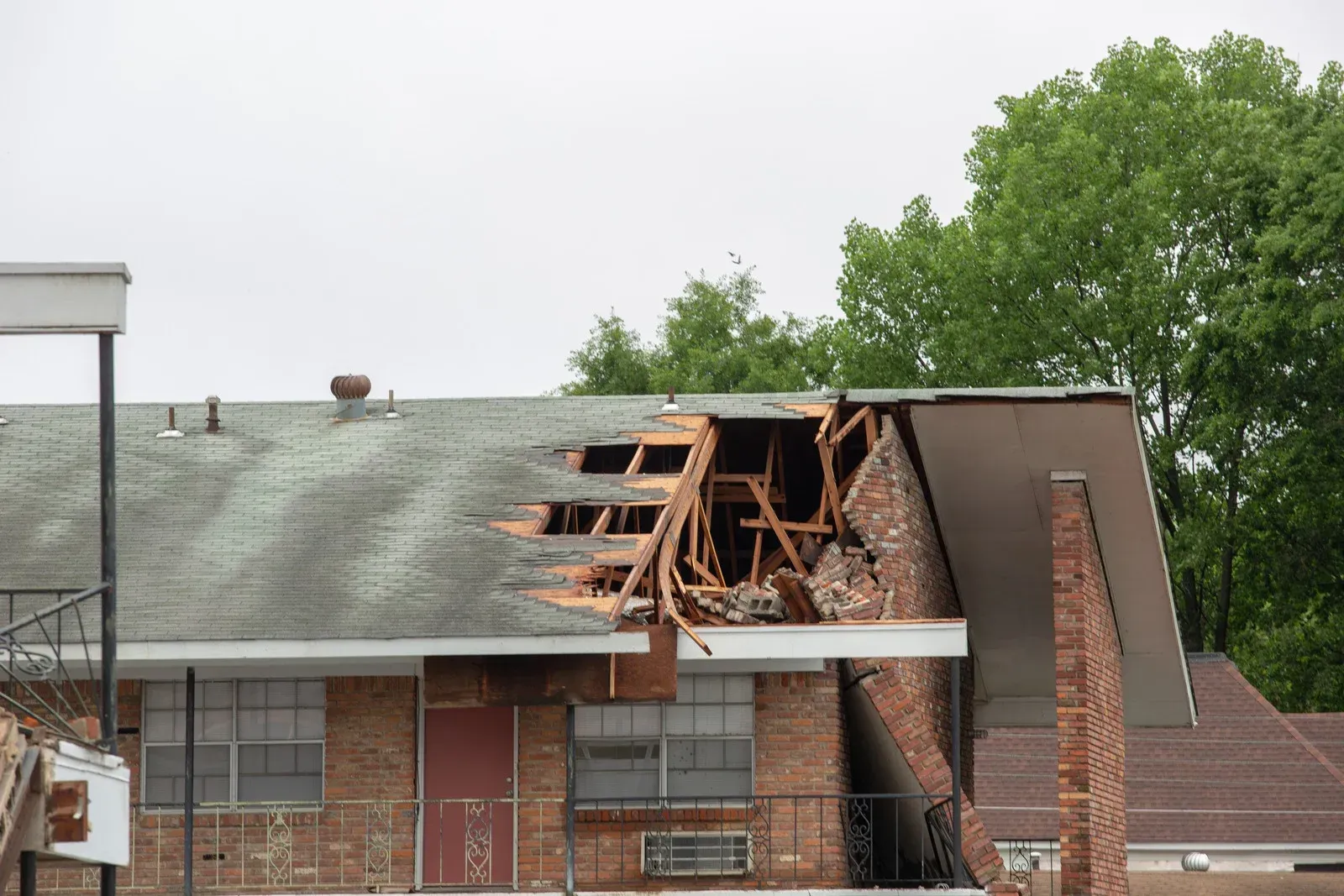 A brick apartment building with significant damage to the roof and an upper wall, revealing exposed wooden support beams.