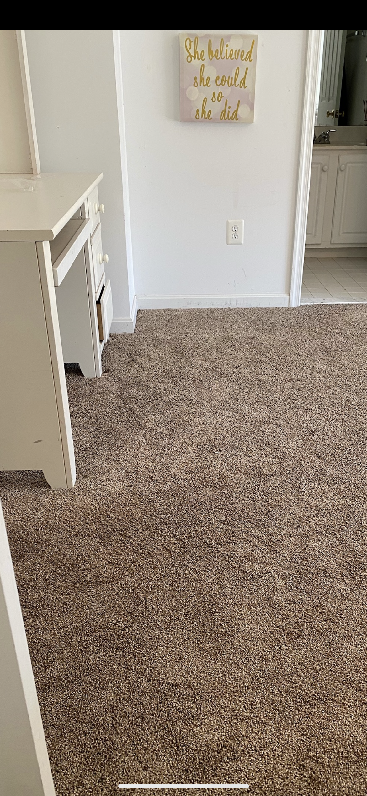 A room with white desk, doorway, and speckled brown carpet. A framed sign hangs on the wall.