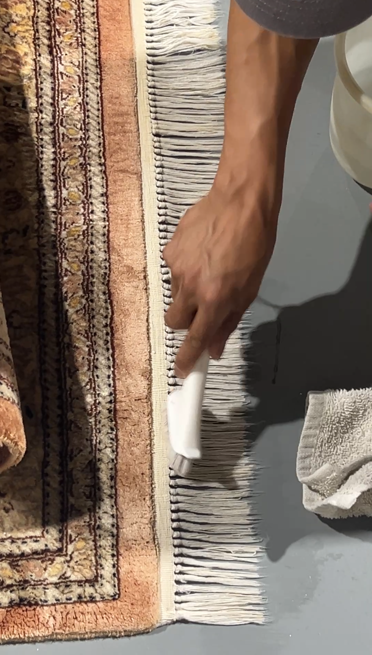 Person cleaning rug fringe with a brush and bucket nearby on a concrete floor.