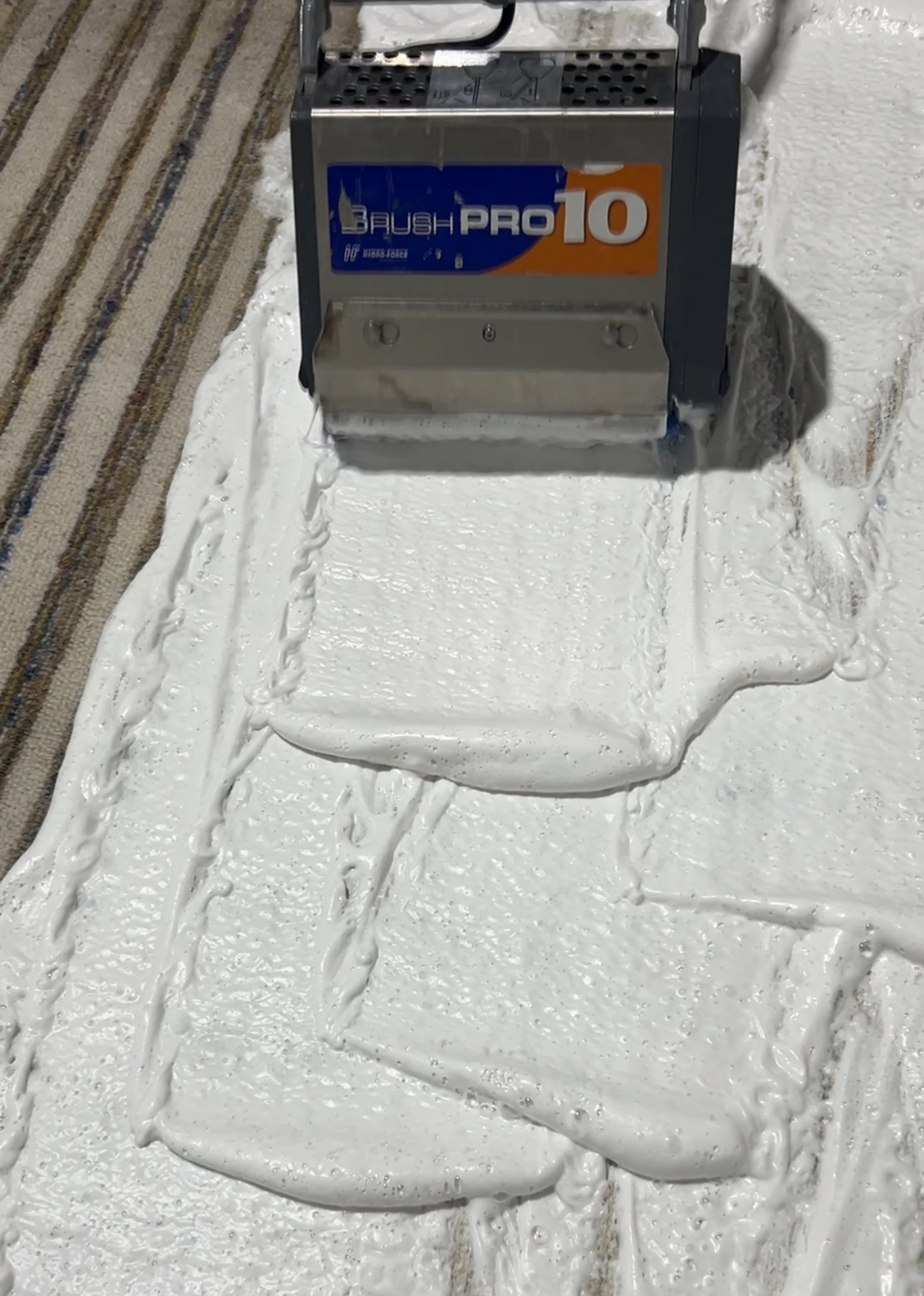 A machine spreading white sealant on a patterned carpet. The machine is labeled