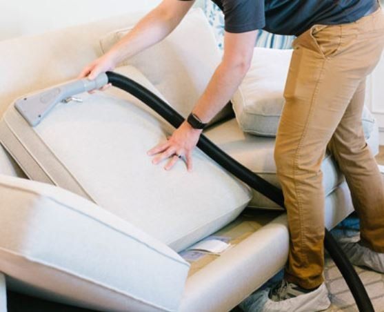 Person vacuuming a cream-colored couch cushion. They are wearing a black shirt and khaki pants.