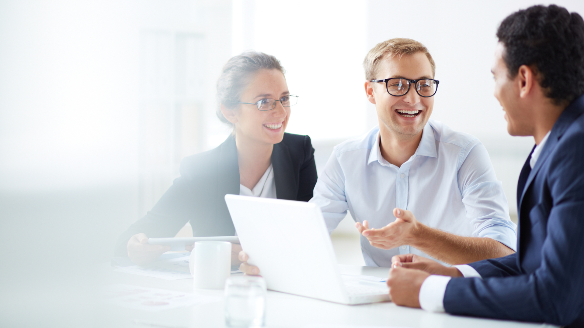Three people in business attire, smiling and talking around a laptop in a bright office setting.