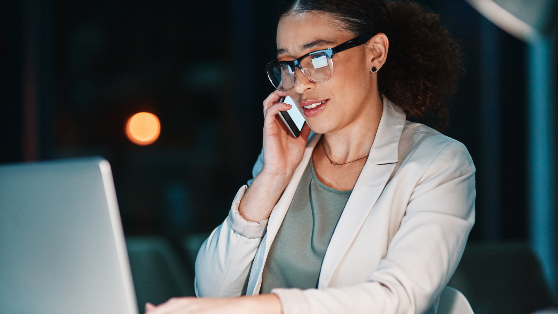 Woman in glasses, jacket, on phone, working at a laptop in a dimly lit office at night.