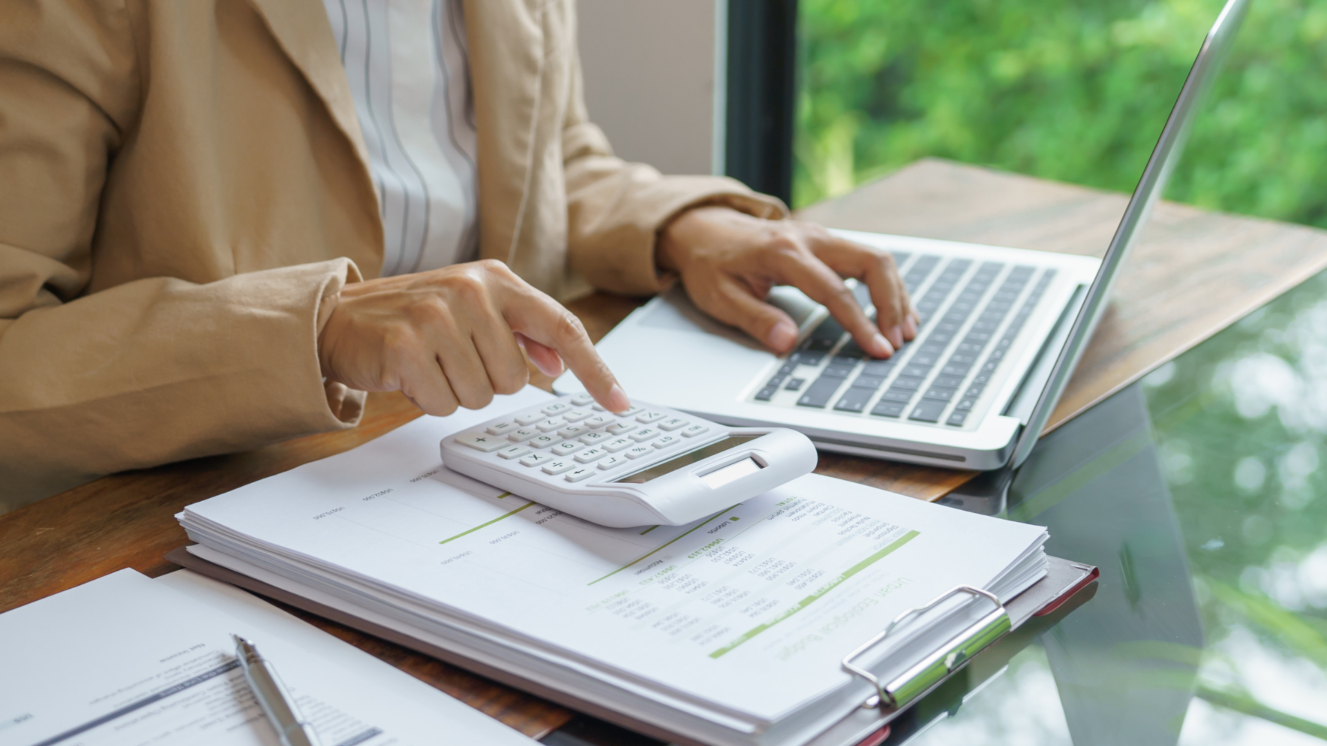 Person using a calculator and laptop with documents on a desk, near a window.