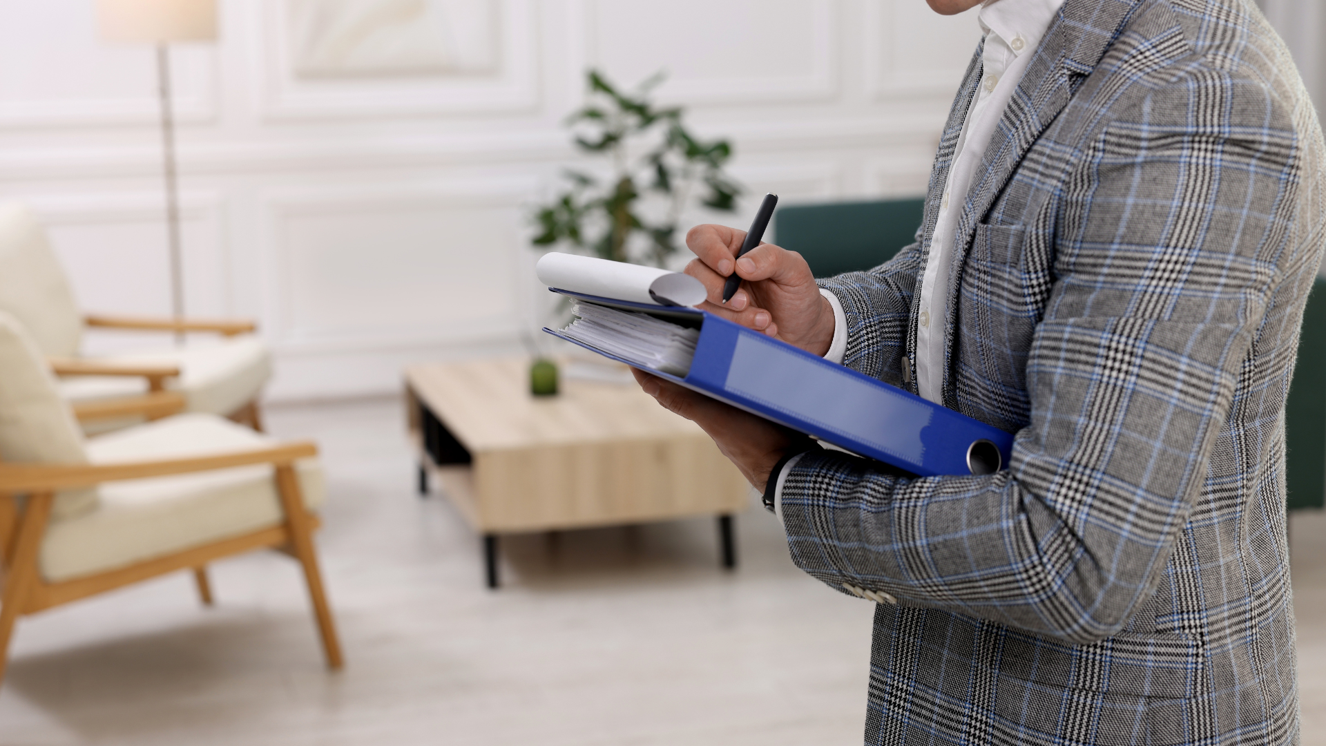 Person in a plaid blazer writes on documents in a blue binder in a bright room.