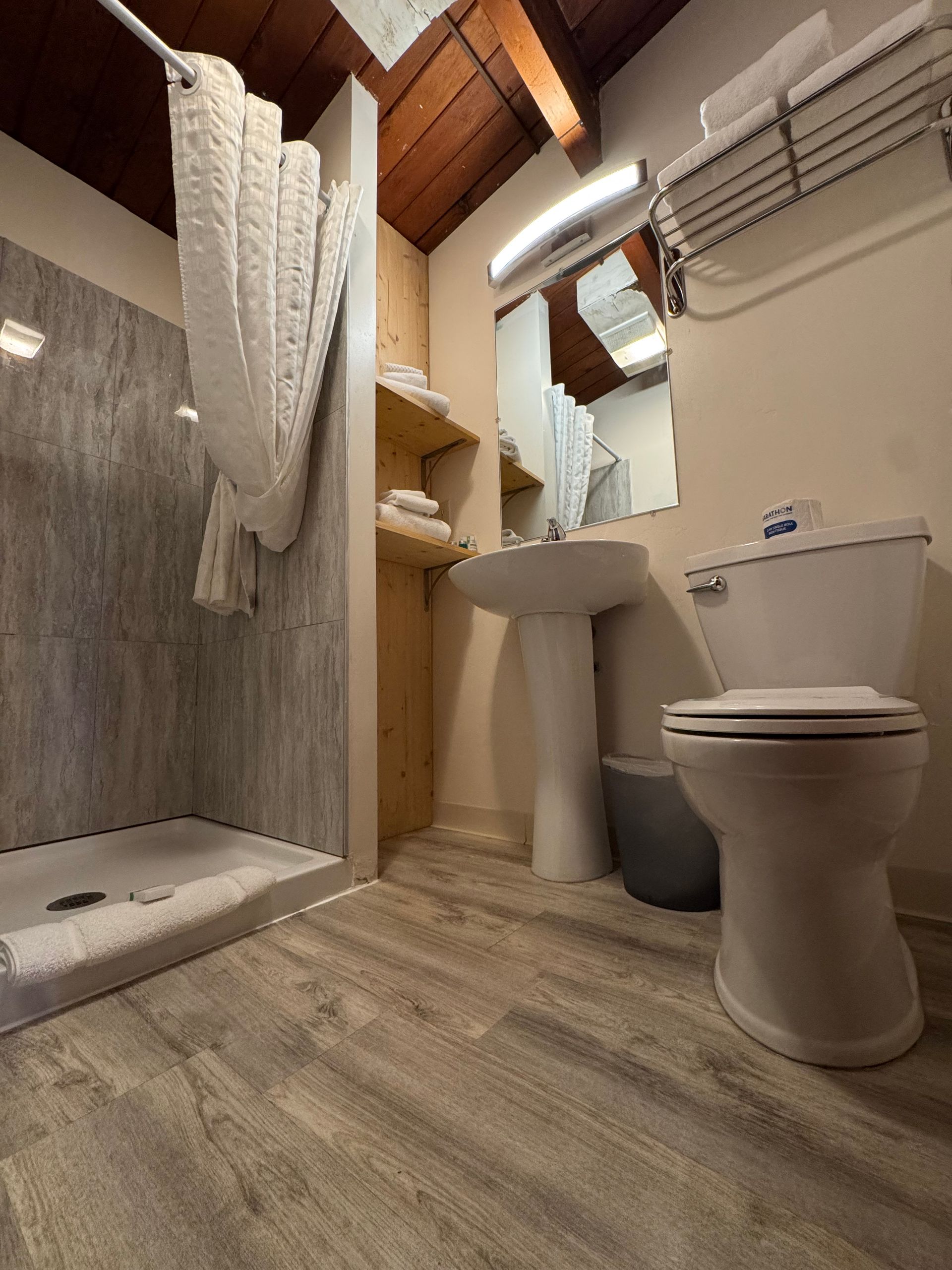 Bathroom with a shower, toilet, sink, and shelves. Light wood floors and an angled ceiling.