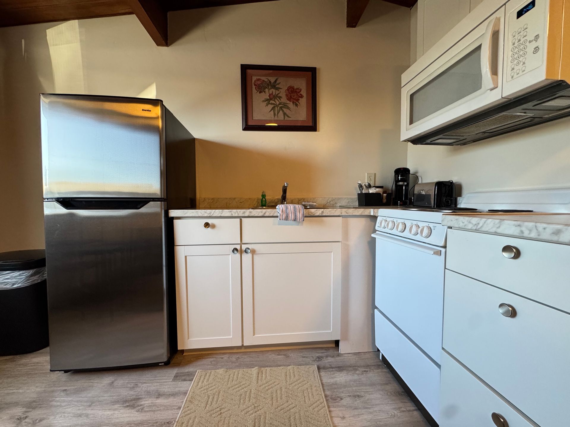 Small kitchen with stainless fridge, white cabinets, stove, microwave, and framed floral art.