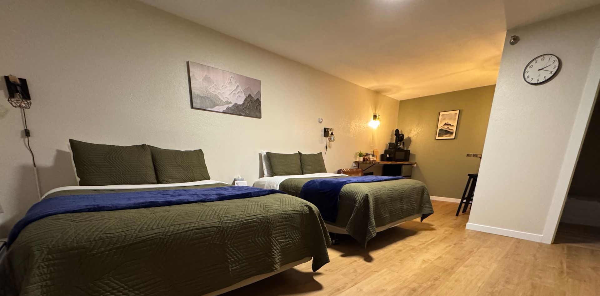A hotel room with two beds covered in olive green quilts, a desk, and a clock on the wall.