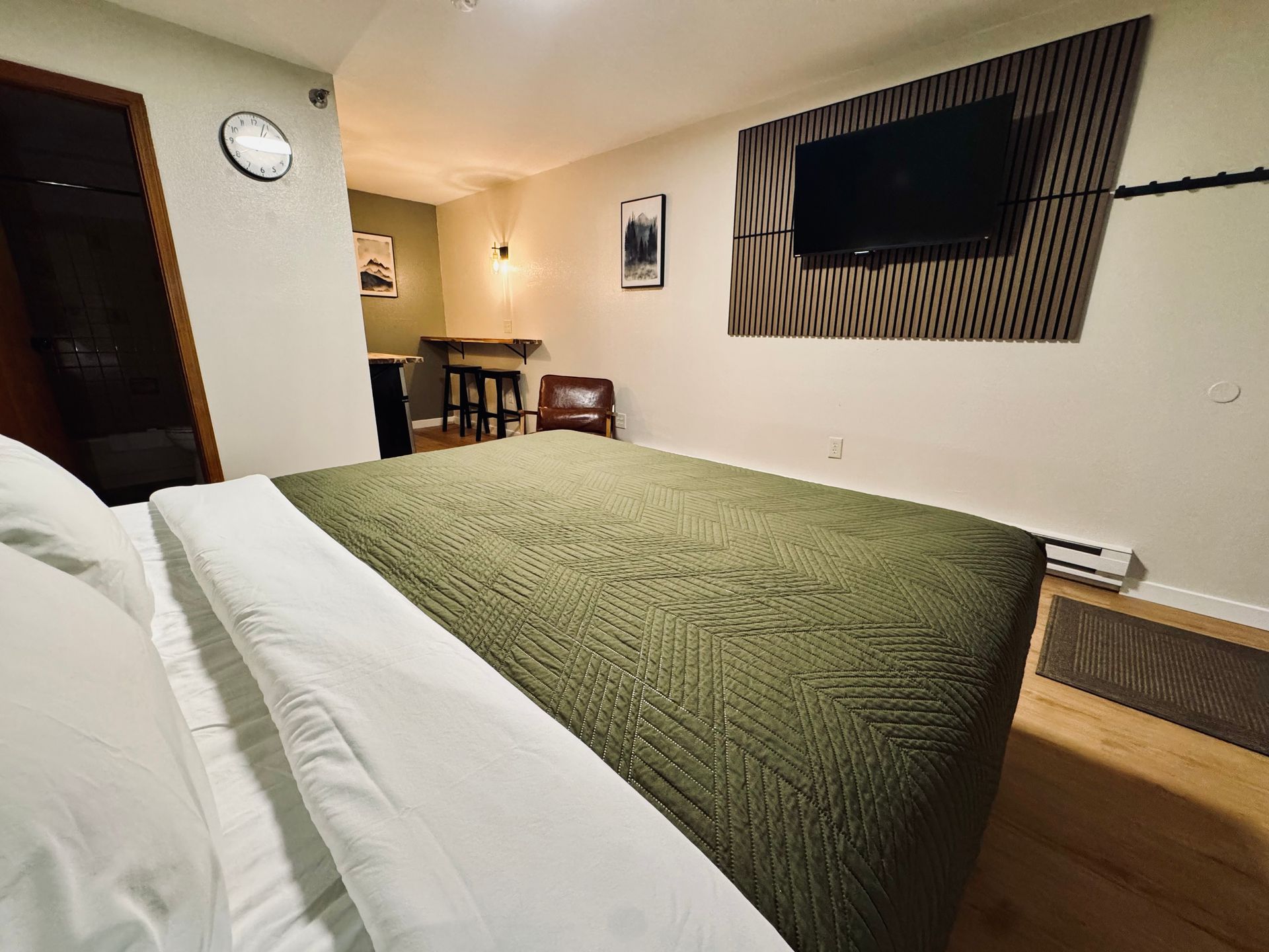 Bedroom with olive green bedspread, TV on textured wall, and small dining area.