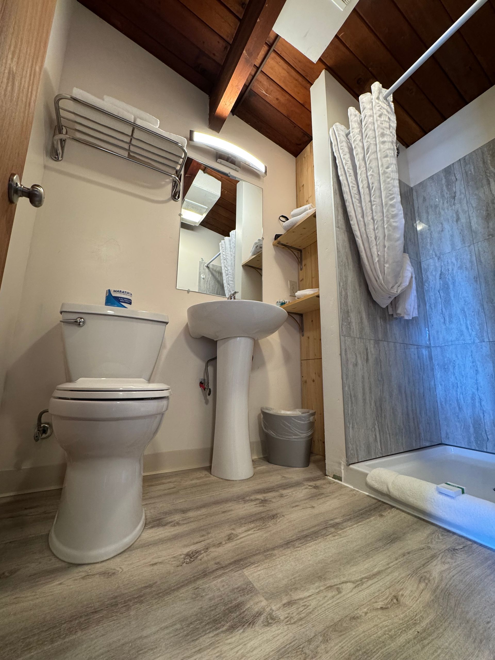 Small bathroom with toilet, sink, and shower; light wood floor, shelves, and ceiling; a neutral color scheme.