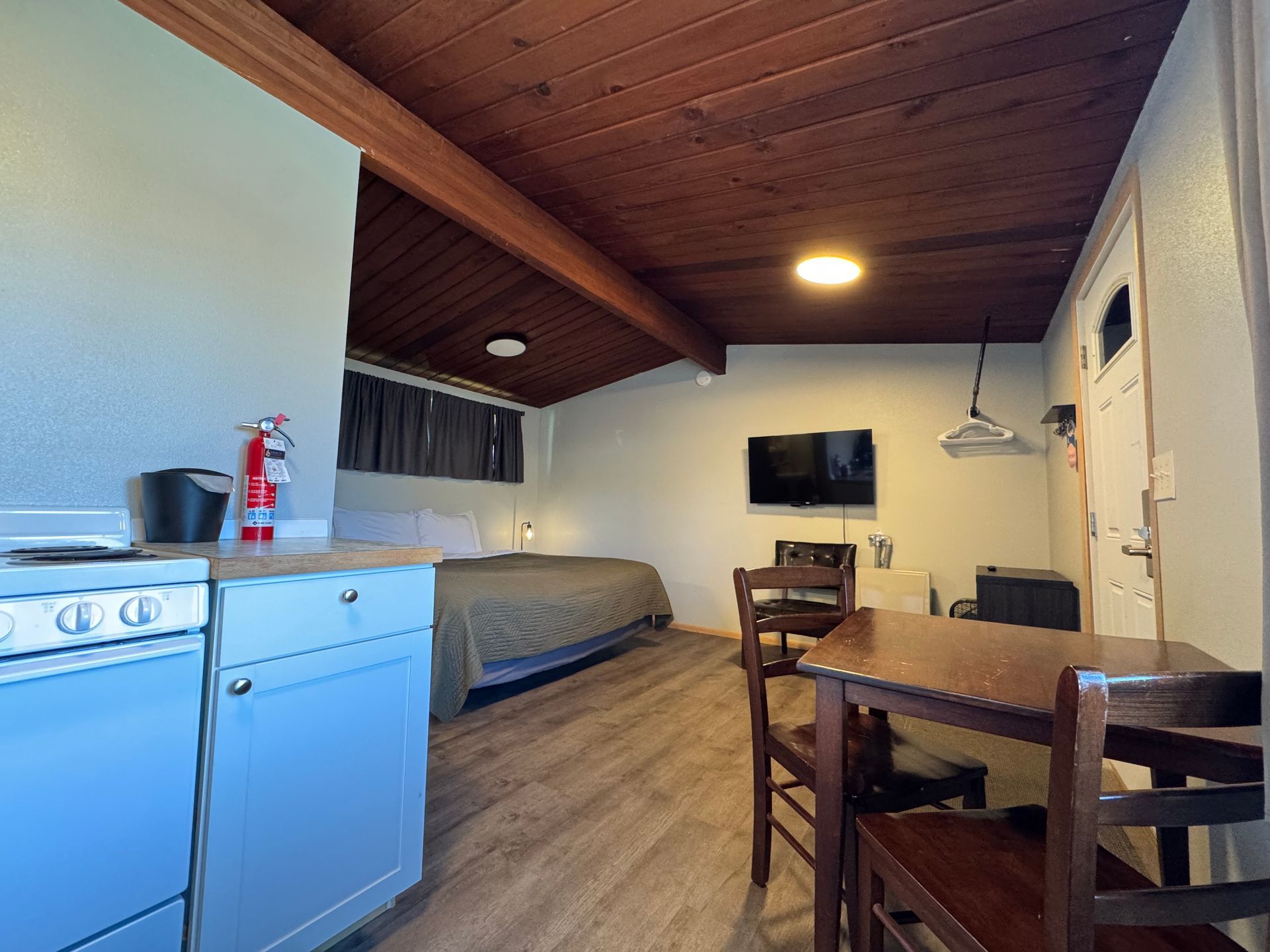 Cozy motel room with bed, table, and small kitchen area. Dark wood ceiling, neutral walls, and hardwood floors.
