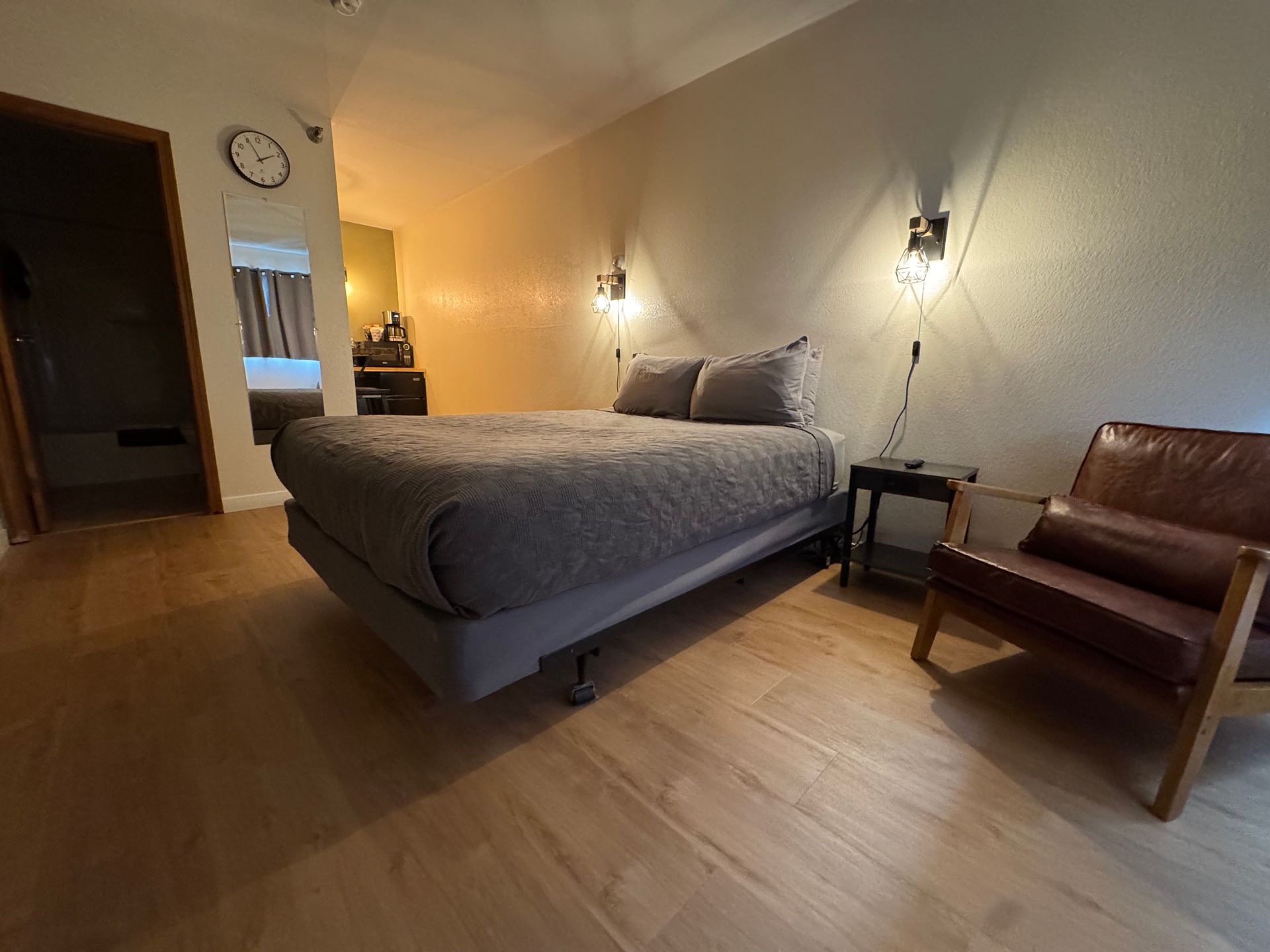 Bedroom with bed, brown chair, and wood floors. Beige walls, two wall sconces, and open doorway.