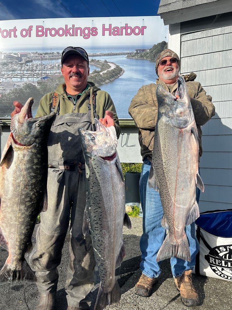Two men are holding large fish in front of a sign that says port of brookings harbor.
