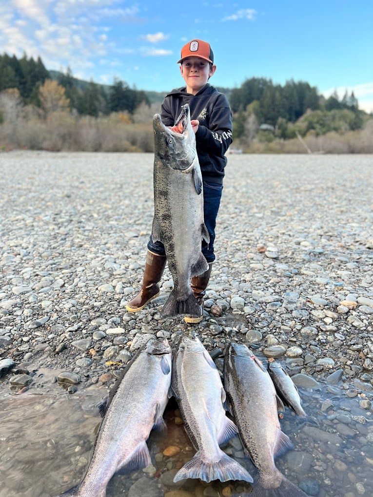 A young boy is standing on a rocky beach holding a large fish.