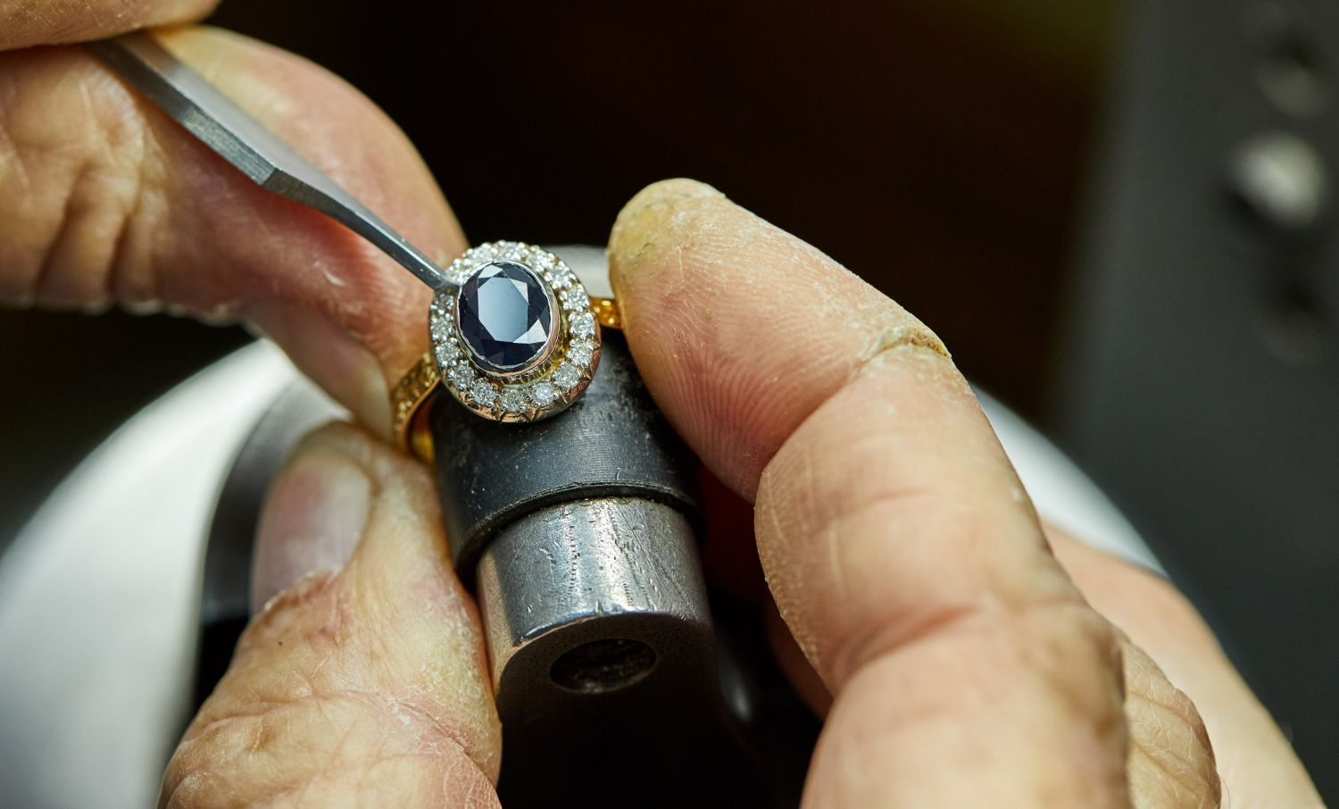 Jeweler’s hands working on a gold ring with a blue gemstone and diamond halo, using tools.