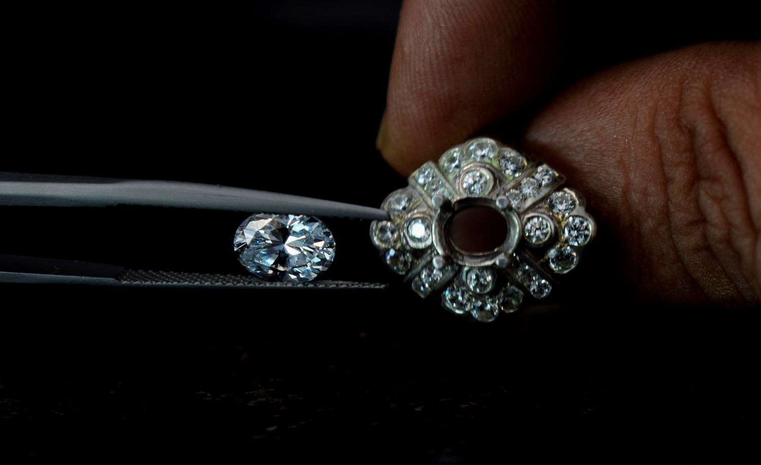 A hand holding a diamond-encrusted jewelry setting with tweezers positioning an oval gemstone to be set into the center.