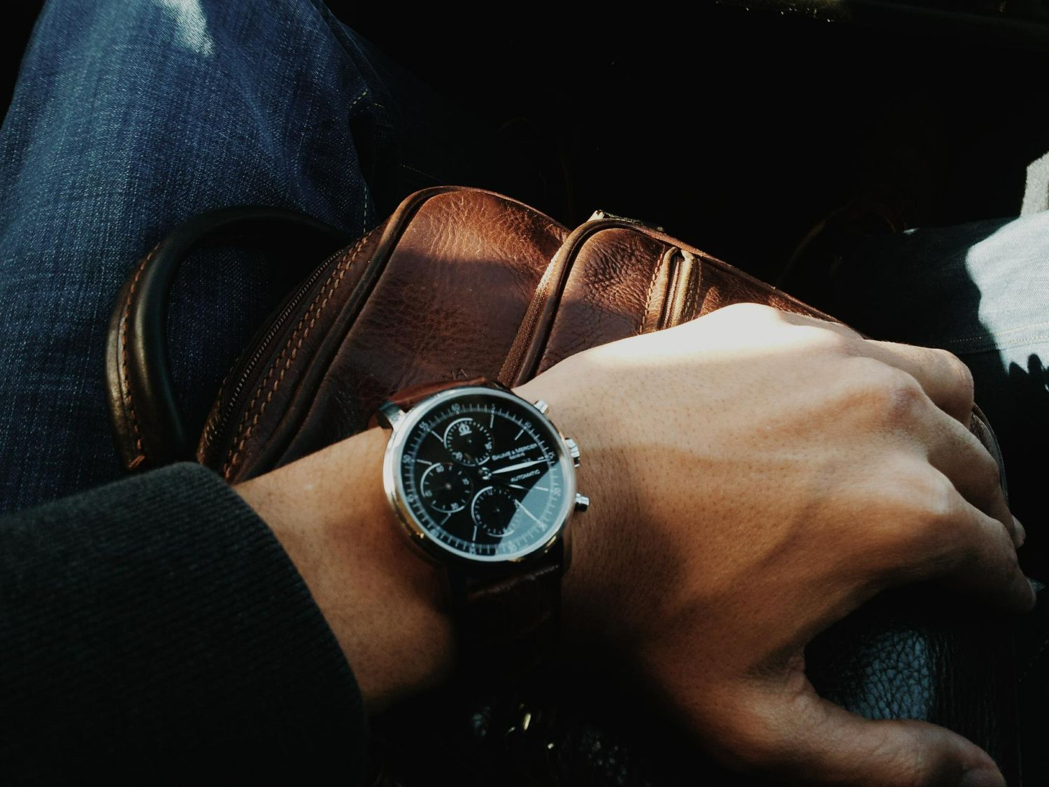 A wristwatch on a person's wrist, brown leather strap, dark face, driving. Brown leather bag visible.