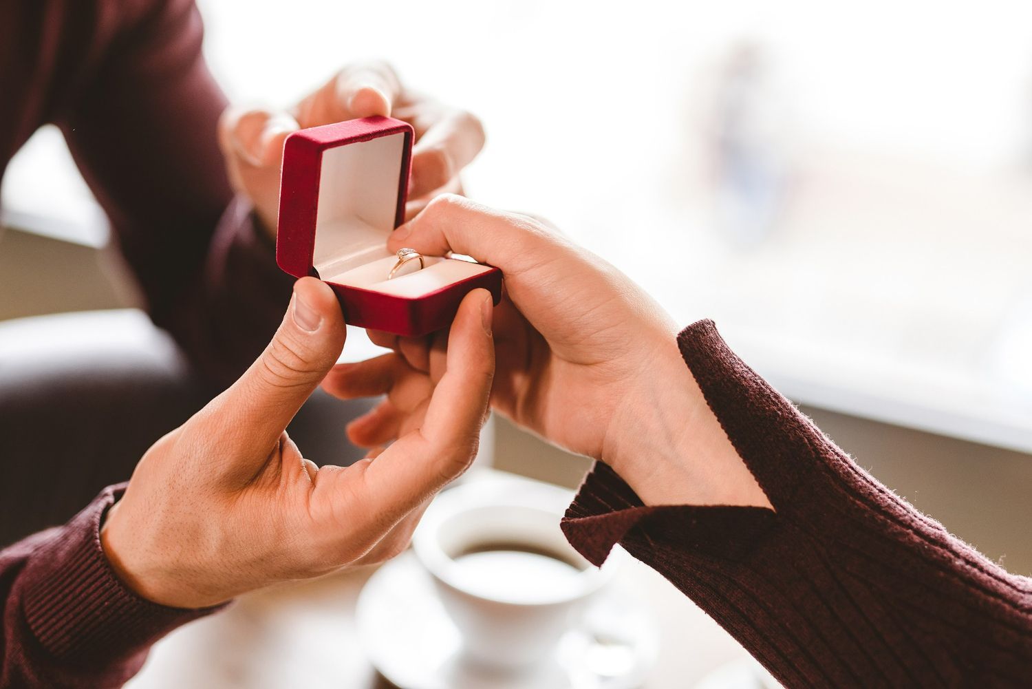 Person offering engagement ring in a red box, near a coffee cup.