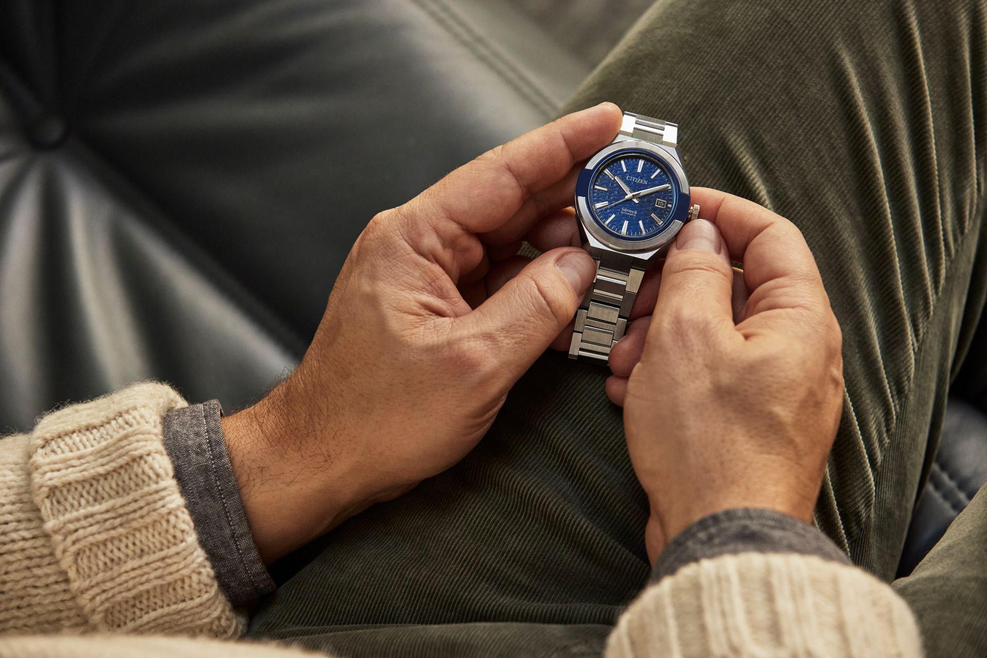 A person wearing a beige sweater and green pants holds a silver wristwatch with a blue dial, adjusting the crown.