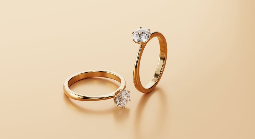Two gold engagement rings with large, clear gemstones on a tan background.