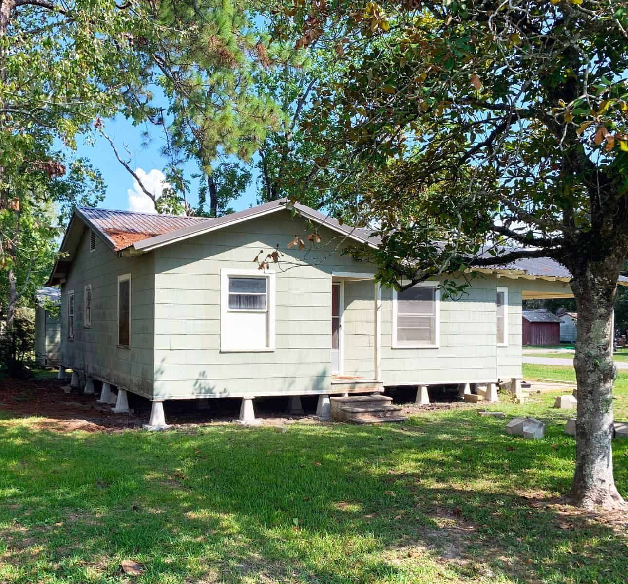 Green house with white trim, set on concrete blocks, under trees.