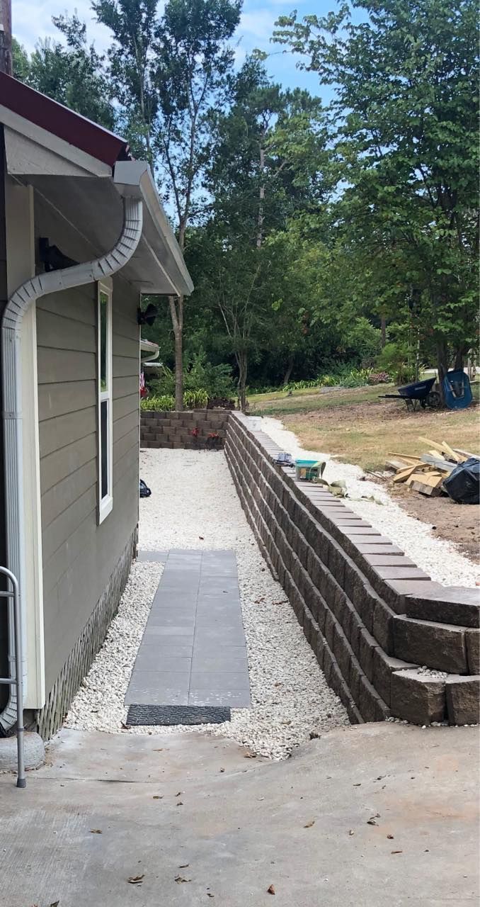 Side of a house with a stone pathway, gravel, and a retaining wall. Green siding, white trim, and trees in the background.