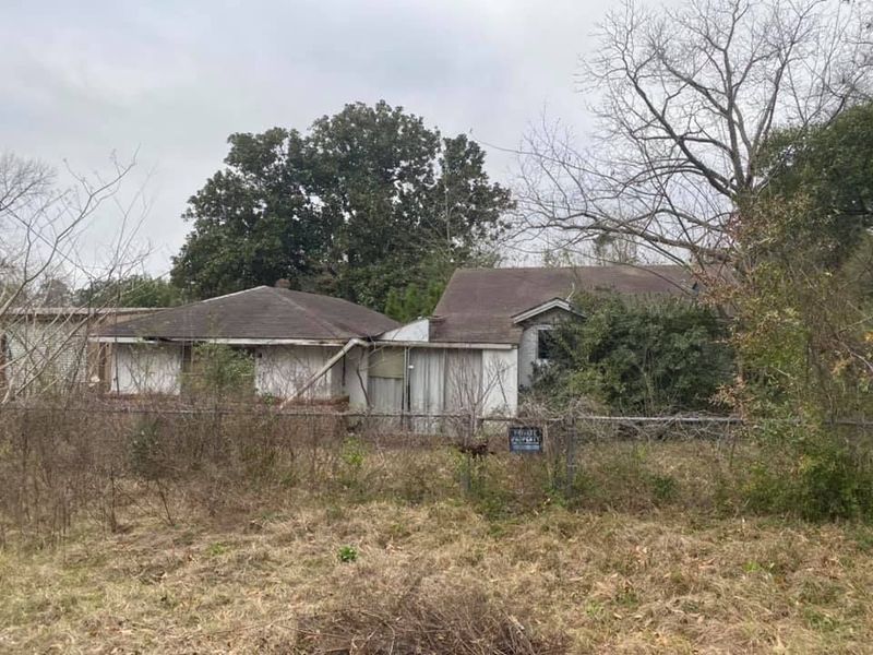 Dilapidated white houses with overgrown vegetation, cloudy sky. 