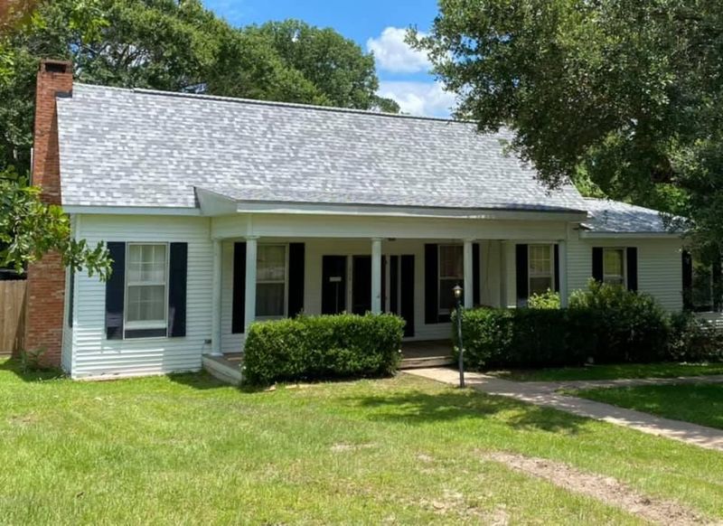 White house with black shutters, chimney, porch, and a gray roof, in a grassy setting.
