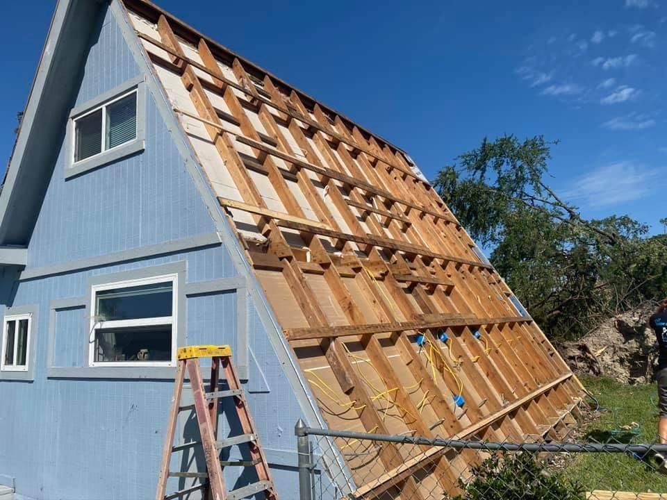 Blue A-frame house with partially removed siding, revealing wooden framing. A ladder leans against the house.