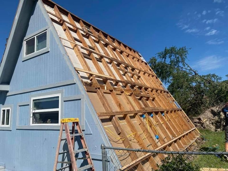 Blue A-frame house with exposed roof beams, a ladder, and a partially removed roof. Sunny day.