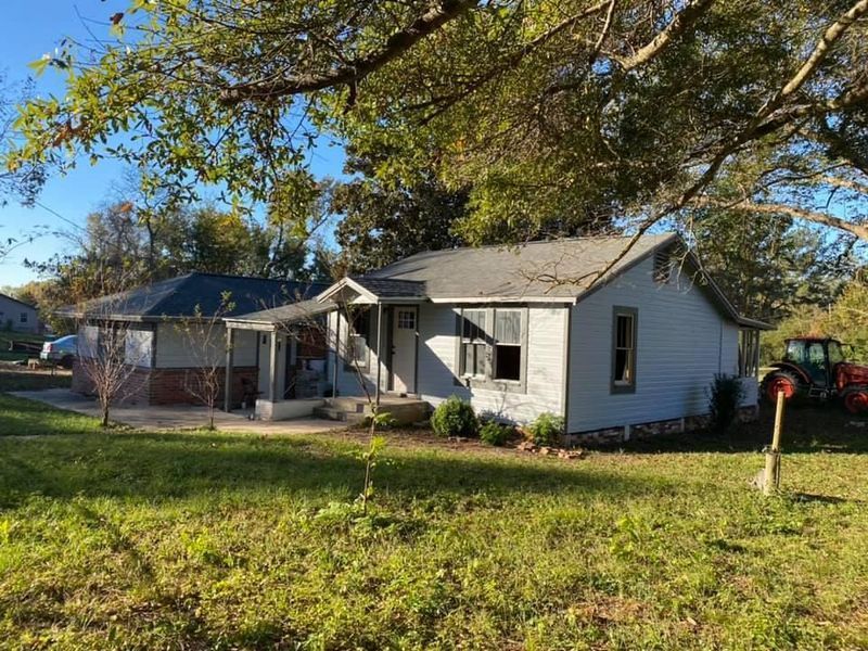 Small, light-blue house with porch and detached carport on a grassy lot under trees; tractor in the background.