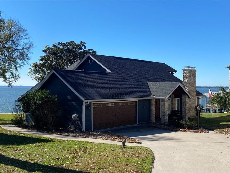 Blue house with dark roof and brown garage door on waterfront. Concrete driveway, chimney, and blue sky.
