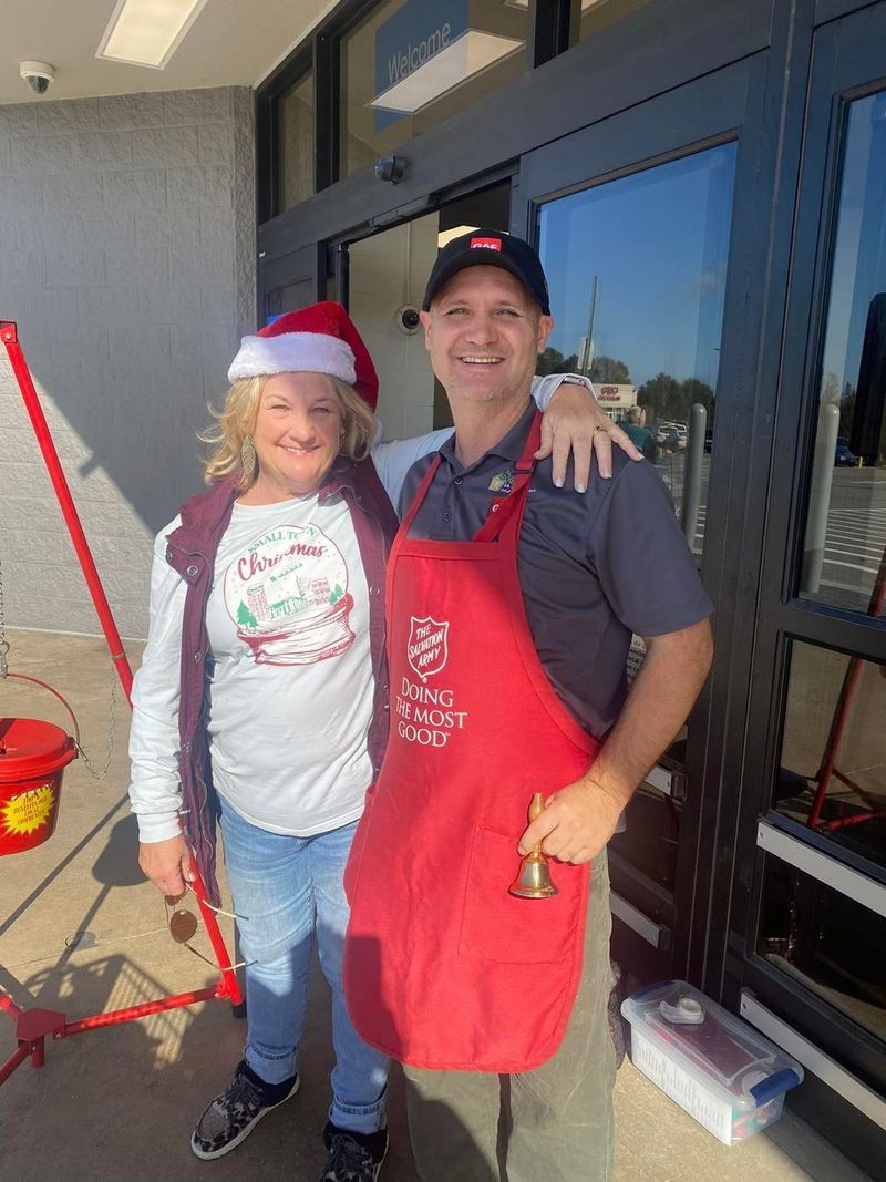 Woman in Santa hat and man in apron pose outside a building, with a Salvation Army kettle nearby.