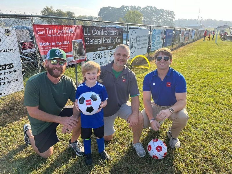 Four people smiling on a soccer field. A child holds a soccer ball. Signage in the background.