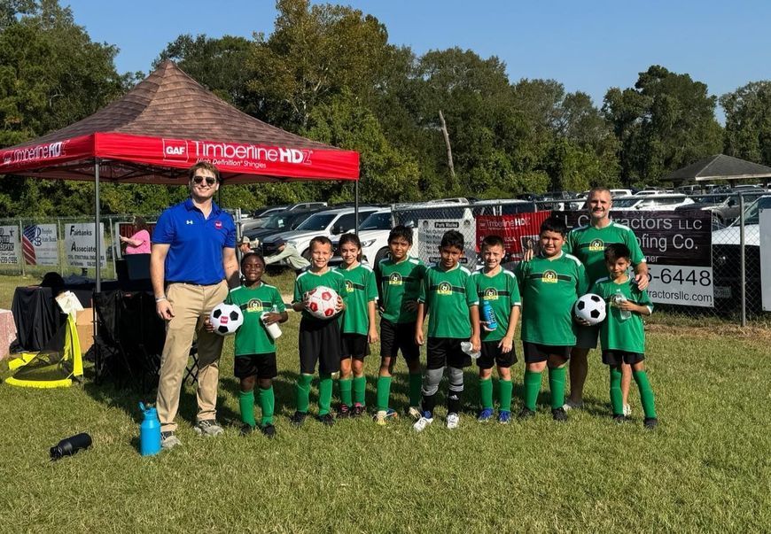 Soccer team in green jerseys poses with coaches in front of a tent and cars on a sunny day.