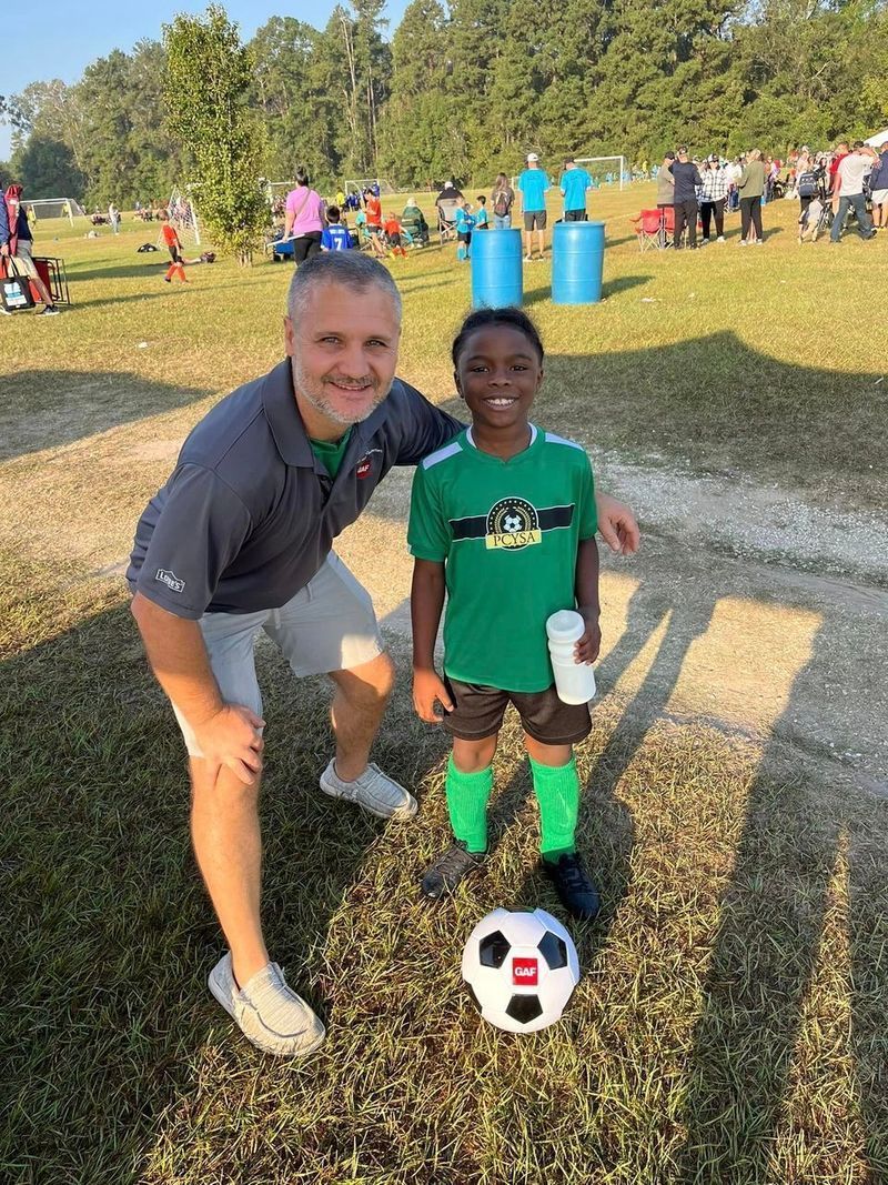 Man and young boy pose for photo on soccer field, ball in front. Boy in green soccer uniform, holding a cup.