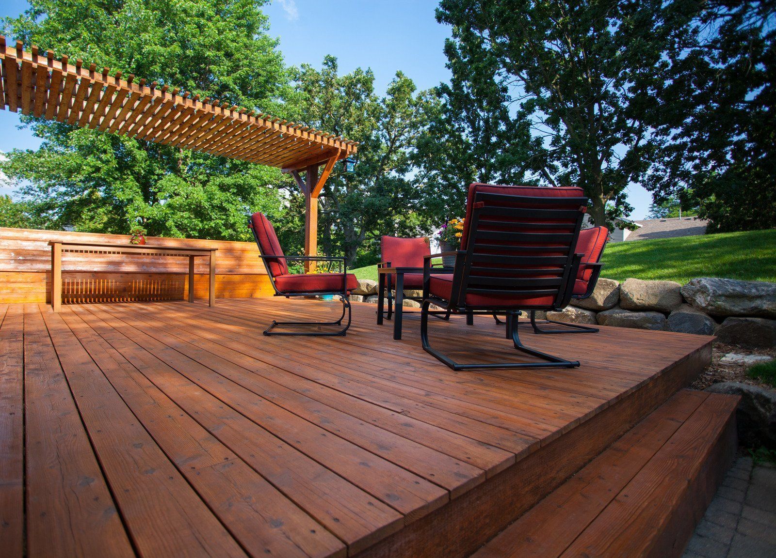 Wooden deck with red chairs under pergola, trees in background.