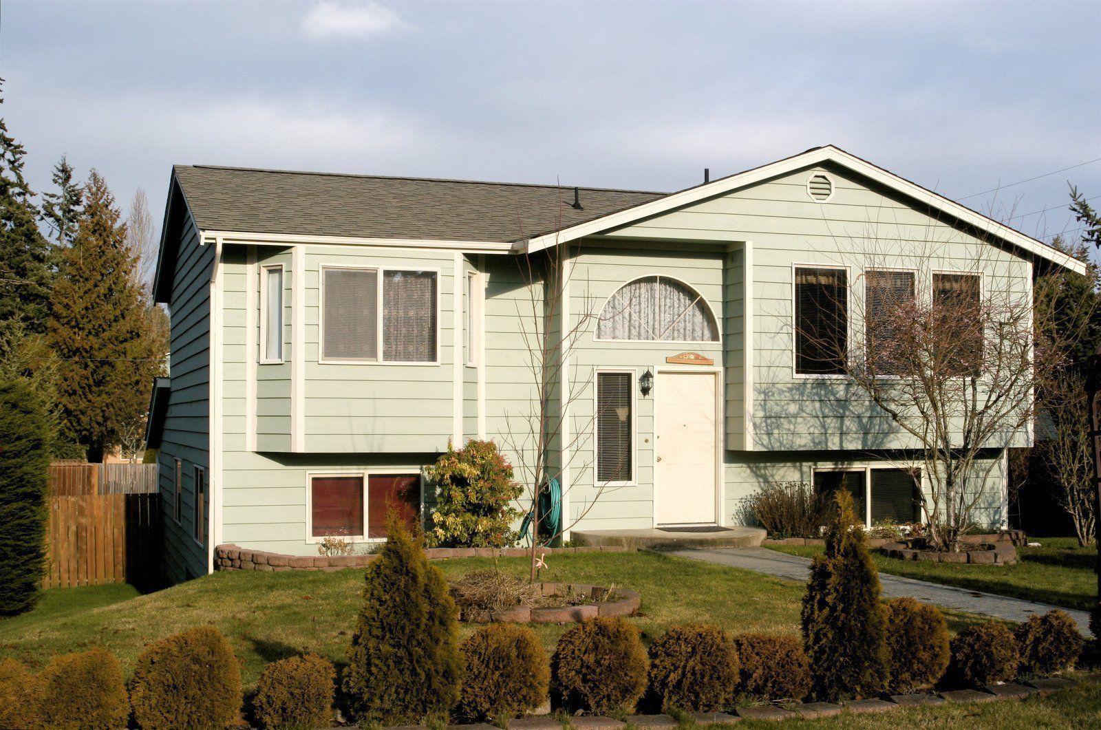 Light green two-story house with a white door, bay windows, and a sloping roof. Lawn and shrubbery in front.