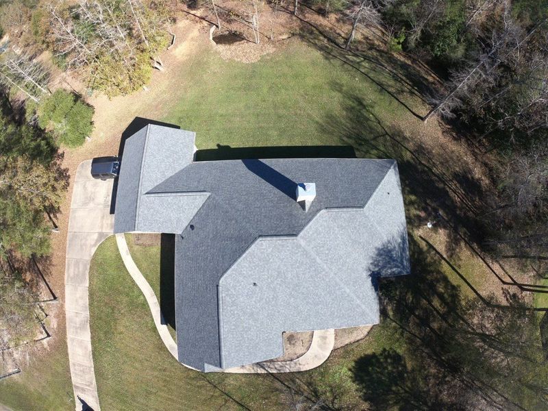 Overhead view of a house with a gray roof, surrounded by green grass and trees. A driveway leads to the house.
