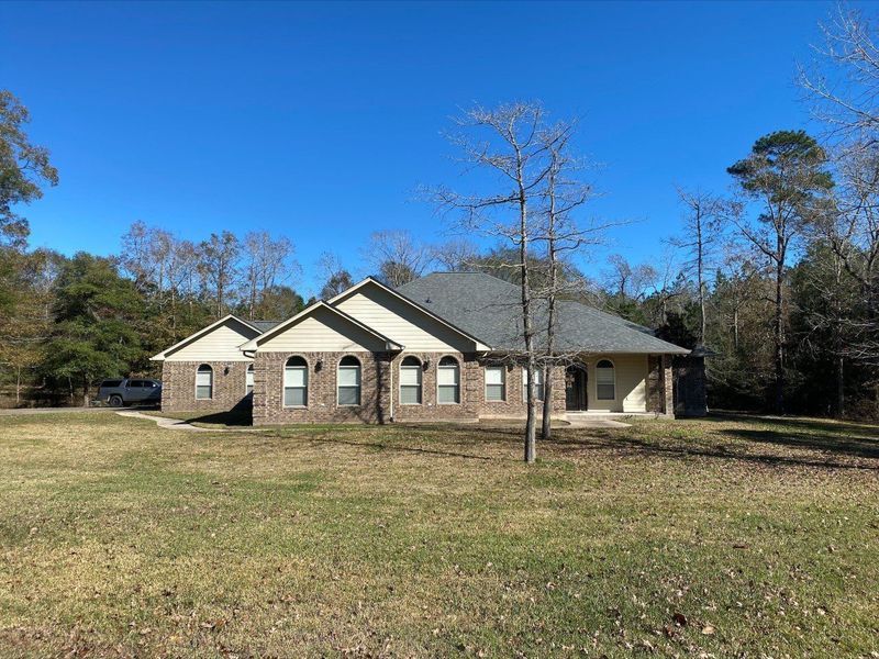 Brick house with dark roof and arched windows on a grassy lot, trees in background, clear blue sky.