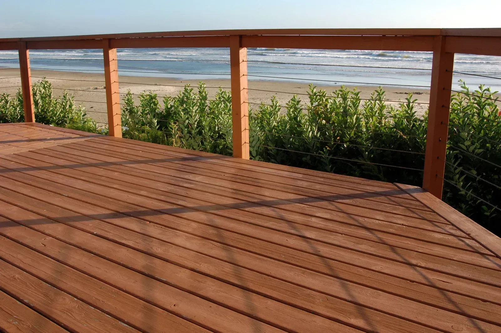 Wooden deck with a beach view, railing, and green foliage. Brown deck boards, blue water.
