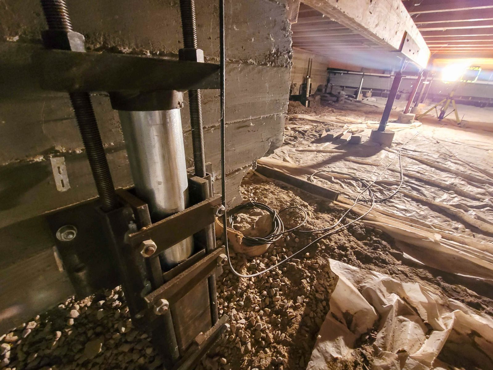 A hydraulic jack supporting a wooden beam in a crawl space filled with dirt and debris.