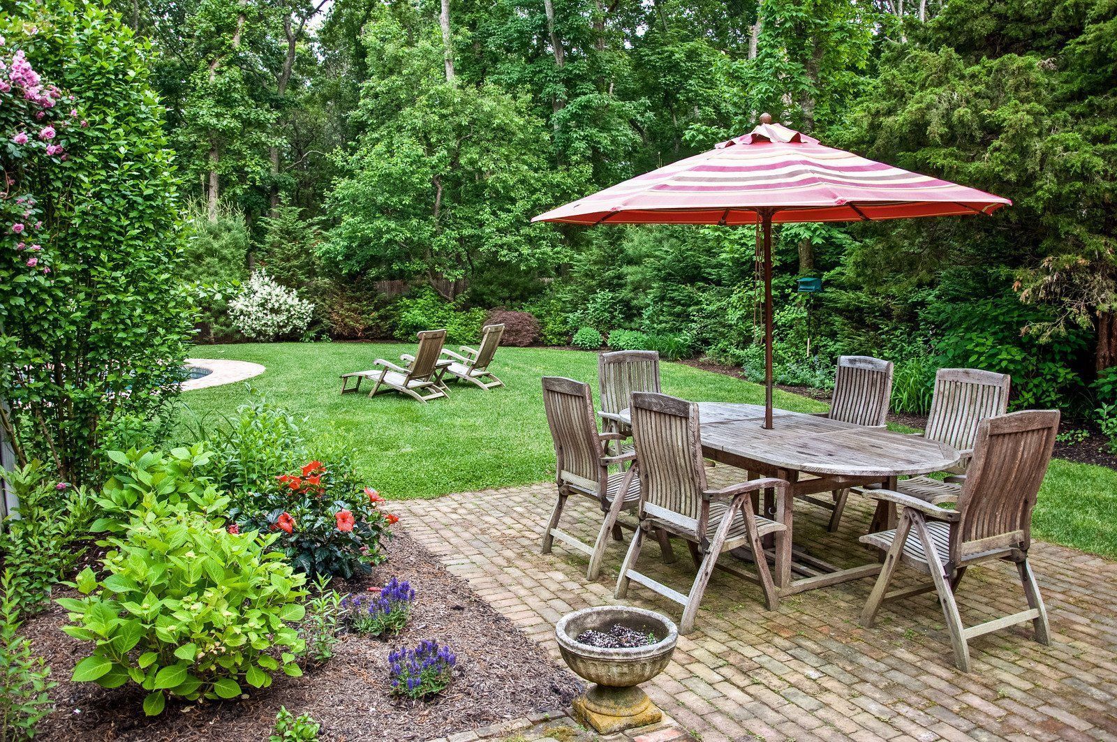 Patio with round wooden table, chairs, and umbrella in lush garden setting.