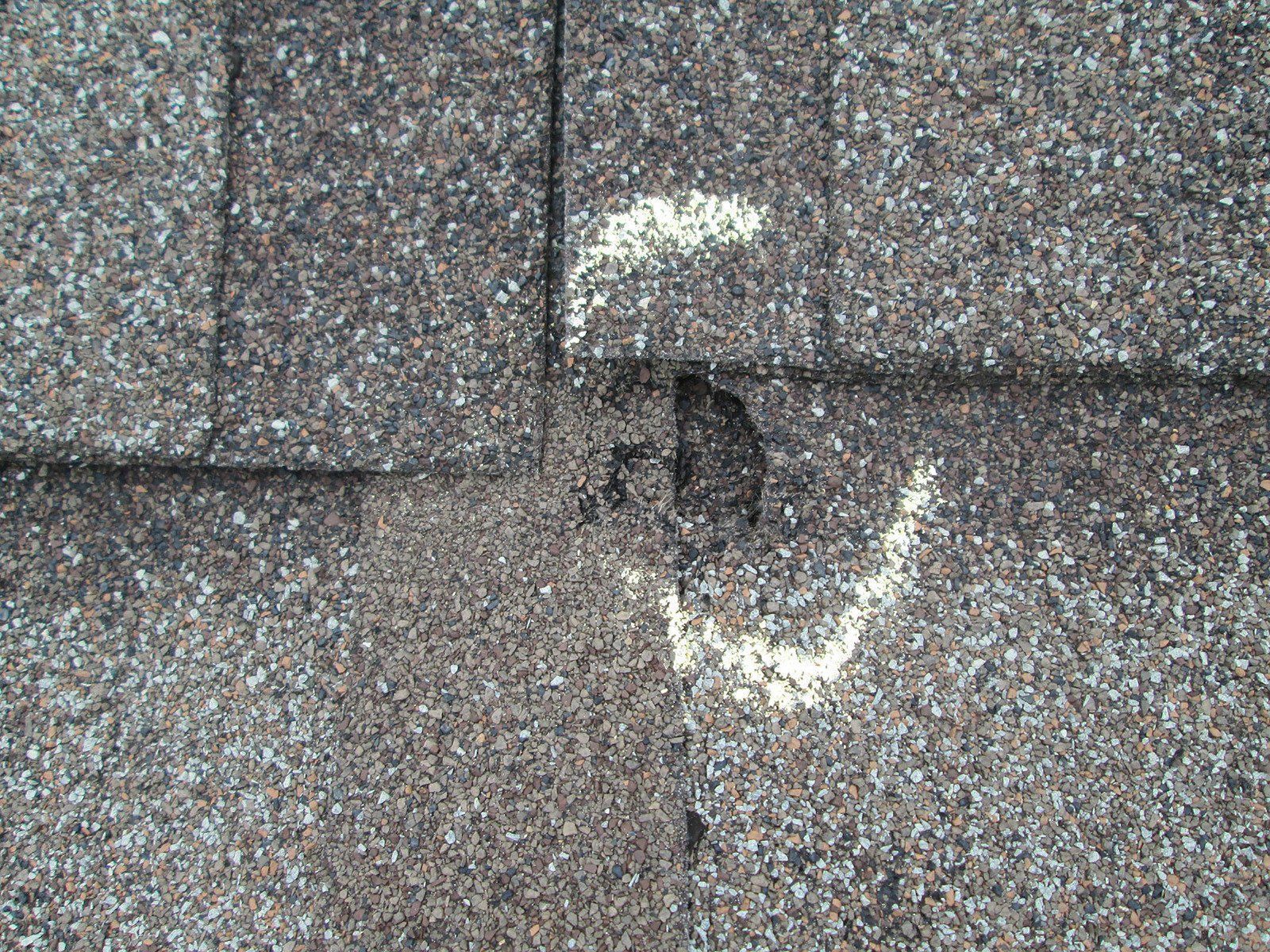 Close-up of asphalt roof shingles with a hole and white markings, likely indicating damage.